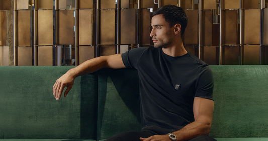 Man wearing a fitted black T-shirt with signature chest badge, seated on a green velvet sofa against a warm gold architectural backdrop.